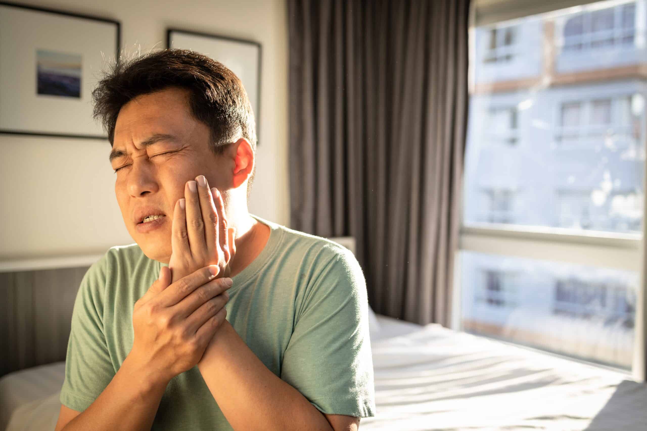 man sitting up in bed holding his jaw, signs of TMJ