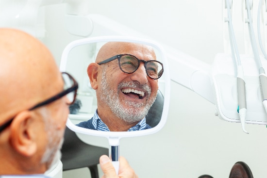 older man looking in a mirror, smiling with his new dental implants