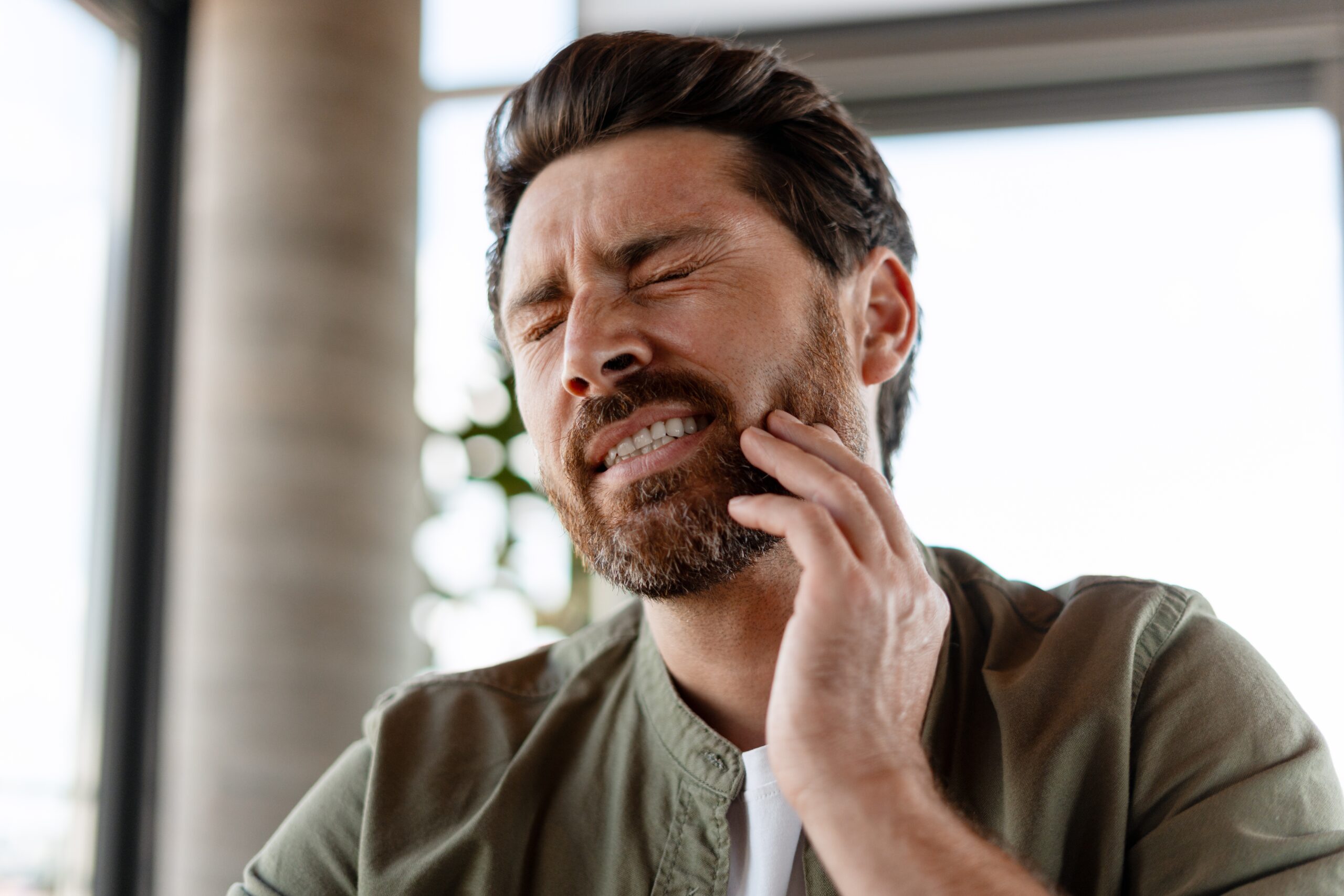 Bearded man experiencing intense tooth and jaw pain, pressing his hand against his cheek in discomfort and distress
